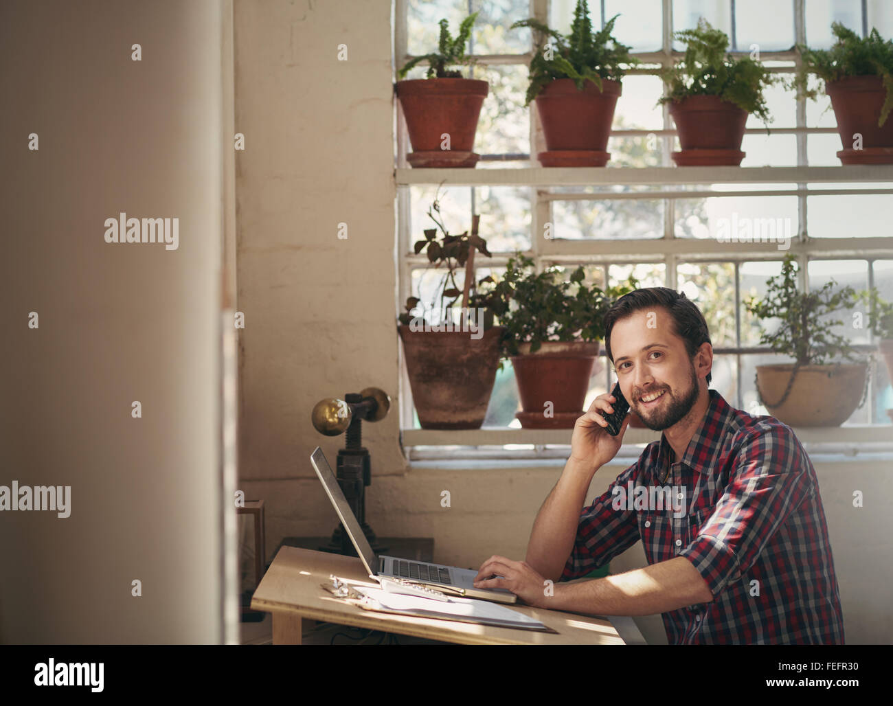 Maschio di designer che lavora presso il suo studio scrivania mentre si parla al telefono e cercando di sorridere alla telecamera Foto Stock