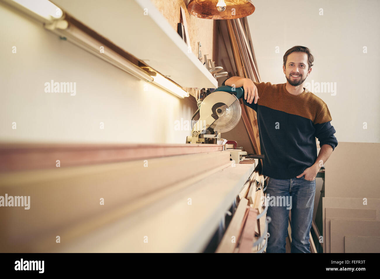 Giovani professionisti corniciaio in posa a fianco di uno strumento specializzato nella sua bottega cercando orgoglioso e sorridente felicemente Foto Stock