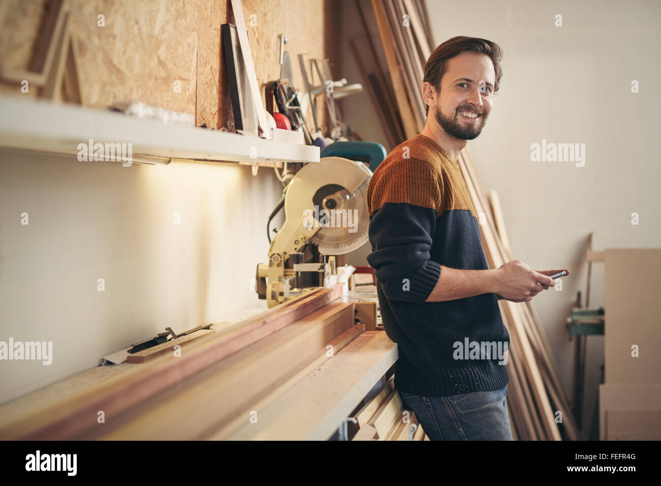 Giovane uomo utilizzando il suo telefono e guardando in alto con un sorriso permanente, mentre casualmente nel suo laboratorio di falegnameria Foto Stock