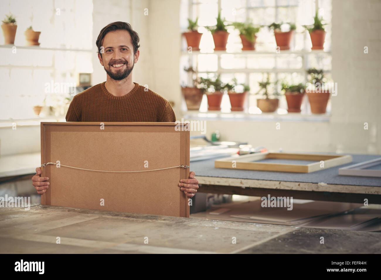 Ritratto di un giovane maschio professional framer tenendo un recente lavoro inquadrato mentre sorridente con fiducia verso la telecamera nel suo studio w Foto Stock