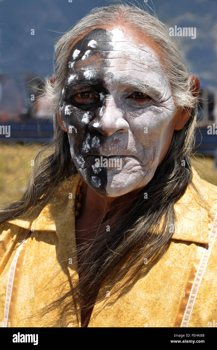 Un volto dipinto Native American sciamano con metà a macchie nere e faccia la metà bianca con lunghi capelli grigi e maglietta gialla Foto Stock
