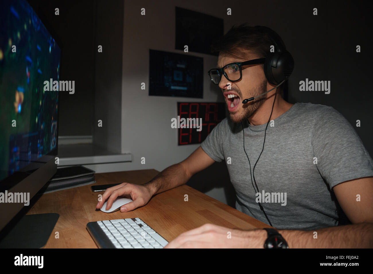 Emozionato bel giovane giocatore in bicchieri e gli auricolari per la riproduzione di giochi per computer e grida a casa Foto Stock