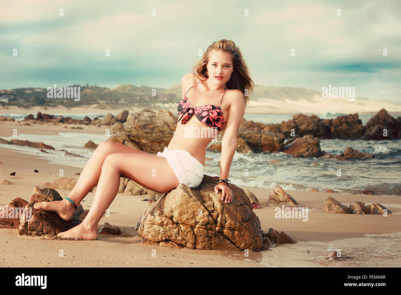 Una bella bionda è seduta su una roccia sulla spiaggia in Africa. Ella ha un bikini bianco e pantaloni corti, la spiaggia può essere anywh Foto Stock