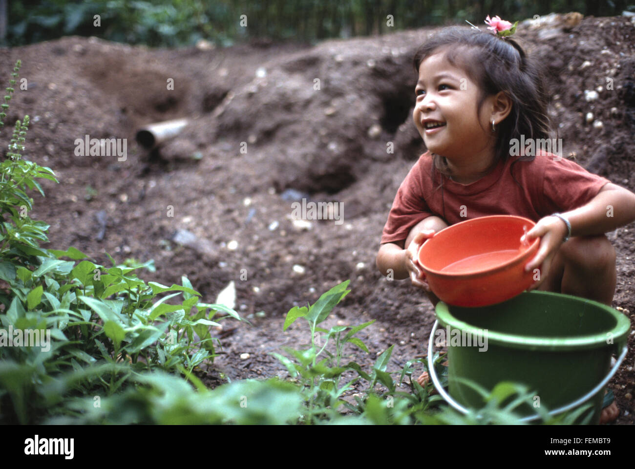 Bambina nel suo giardino contribuendo ad annaffiare le piante e giocando con un secchio di plastica e la vaschetta in un villaggio in Laos Foto Stock