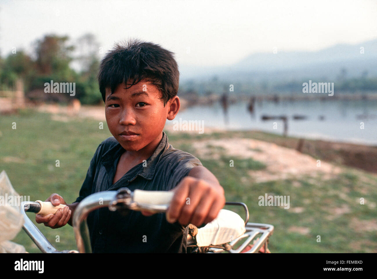Giovane ragazzo sta con la sua bicicletta accanto alla riva del fiume Mekong con il Phou Bia mountain range in background Laos Foto Stock