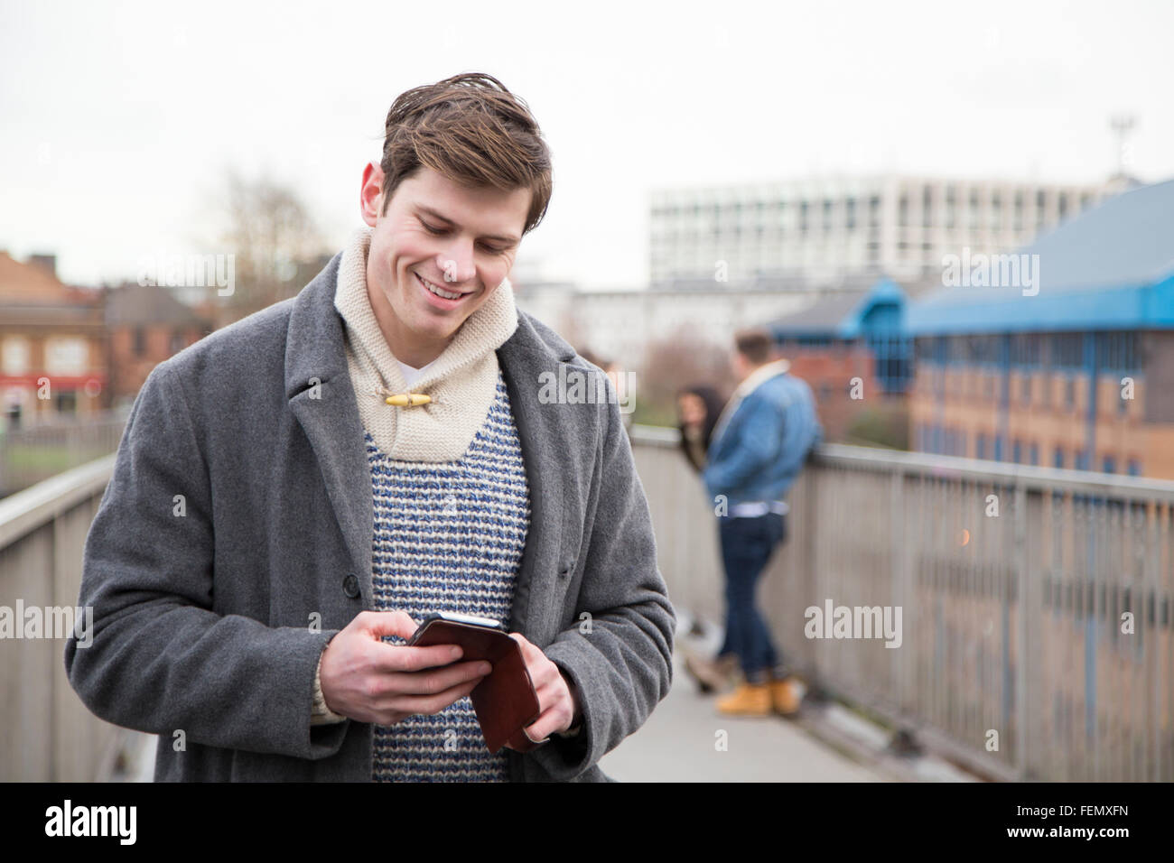 I giovani adulti utilizzando uno smartphone in città. Un gruppo di persone può essere visto a parlare in background. Foto Stock