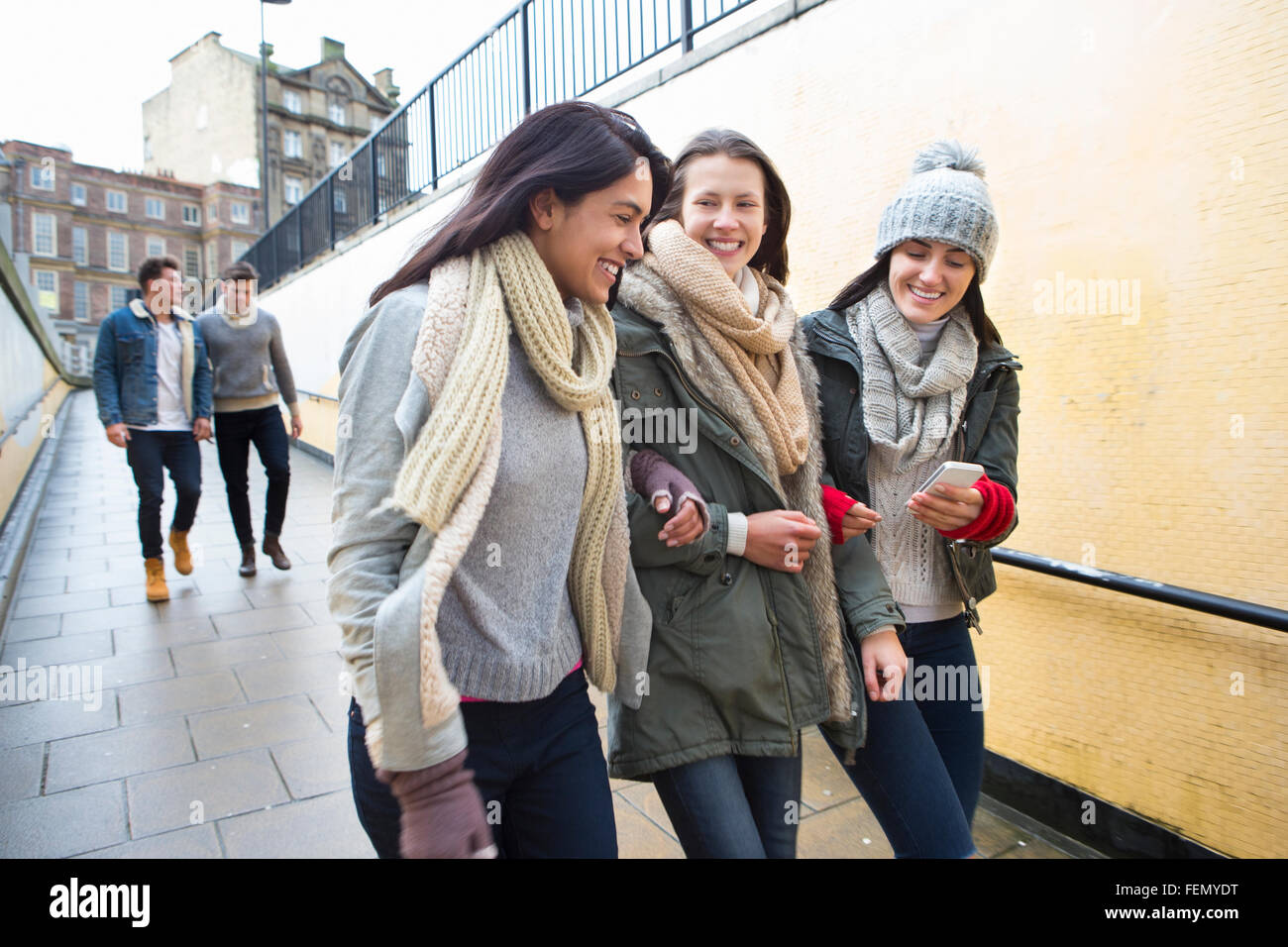 Tre giovani donne a piedi nella città, guardando qualcosa su uno smartphone. Due uomini si può osservare camminando dietro di loro. Foto Stock
