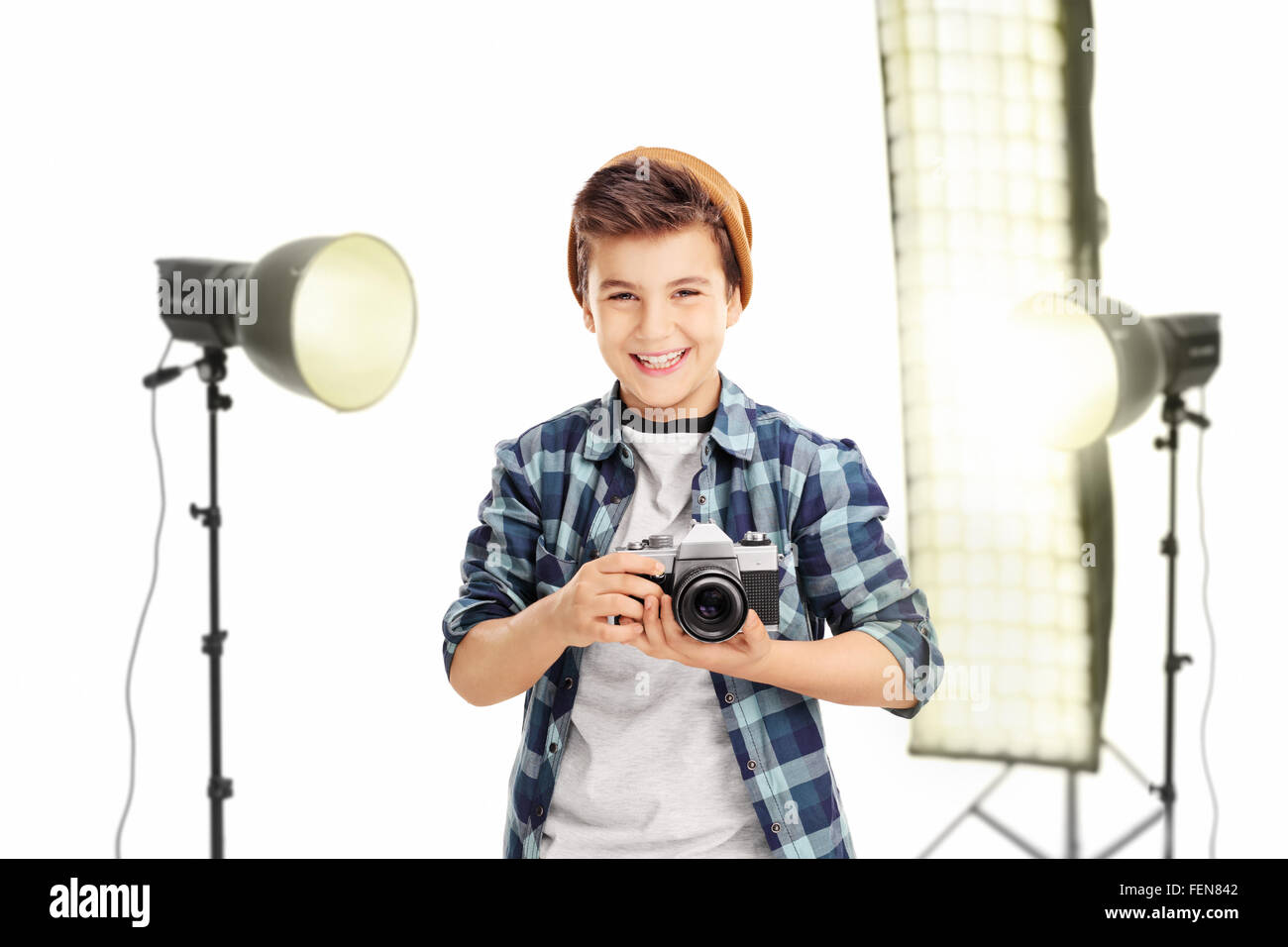 Gioiosa little boy in possesso di una fotocamera e di stare in studio con apparecchiature di illuminazione isolato su sfondo bianco Foto Stock