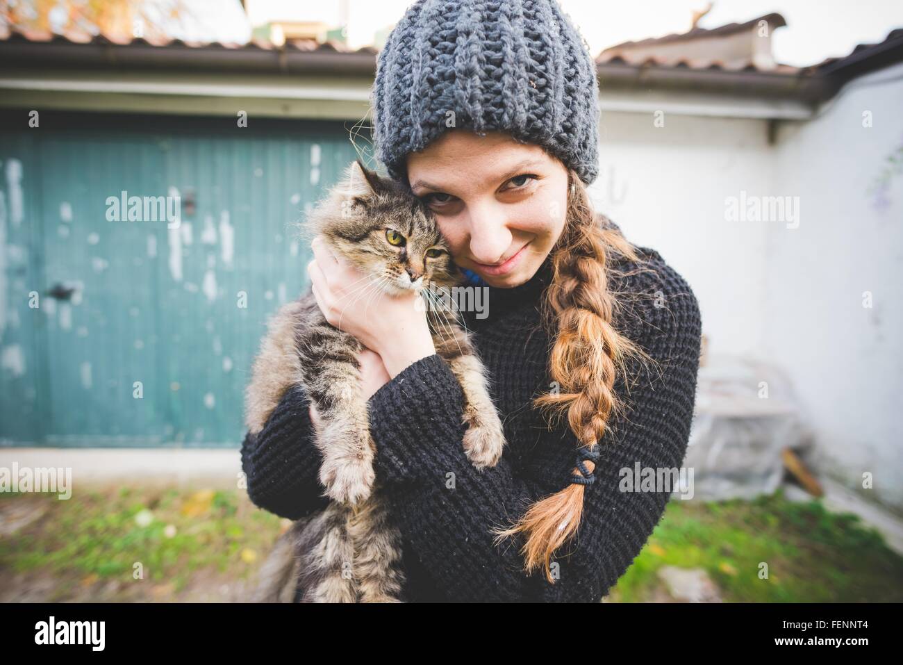 Giovane donna indossa knit hat snuggling cat, guardando alla fotocamera a sorridere Foto Stock