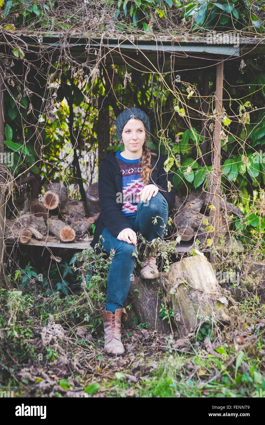 Giovane donna indossa knit hat in seduta di legna da ardere tugurio che guarda lontano Foto Stock