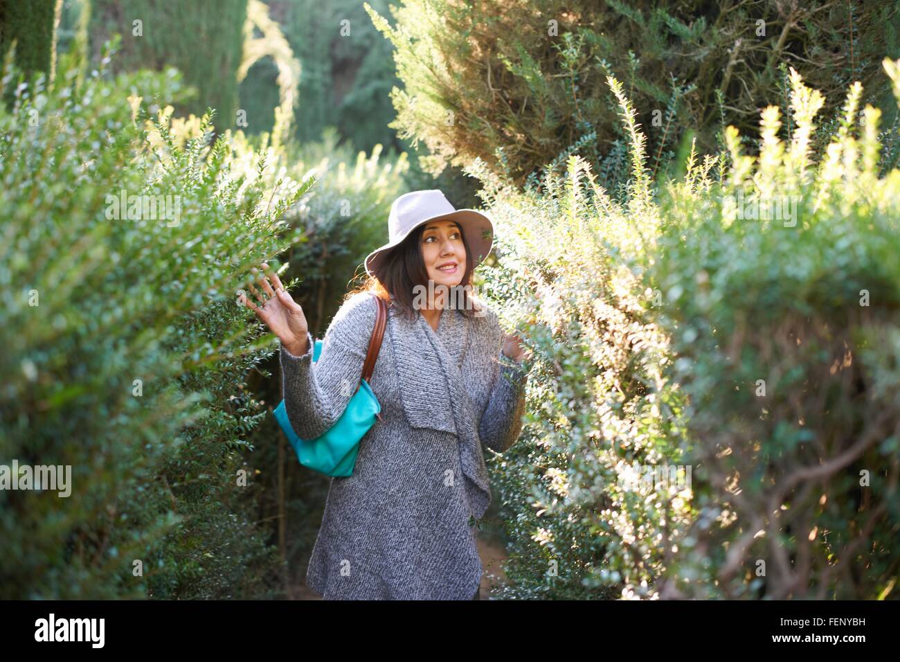 Donna matura indossando cappello e cardigan toccando le piante, guardando lontano sorridente, Siviglia, Spagna Foto Stock