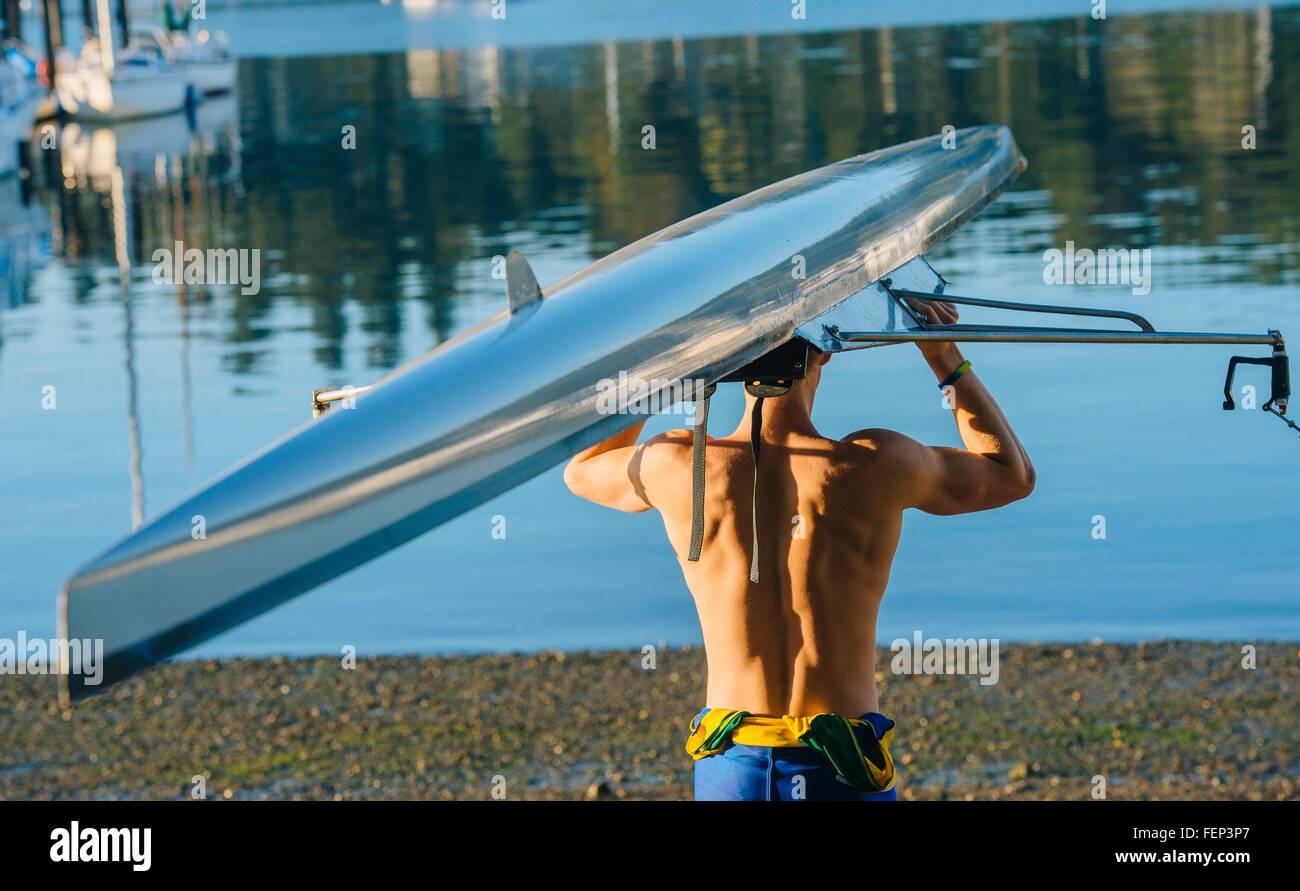 Ragazzo adolescente holding sculling barca al di sopra della testa vista posteriore Foto Stock