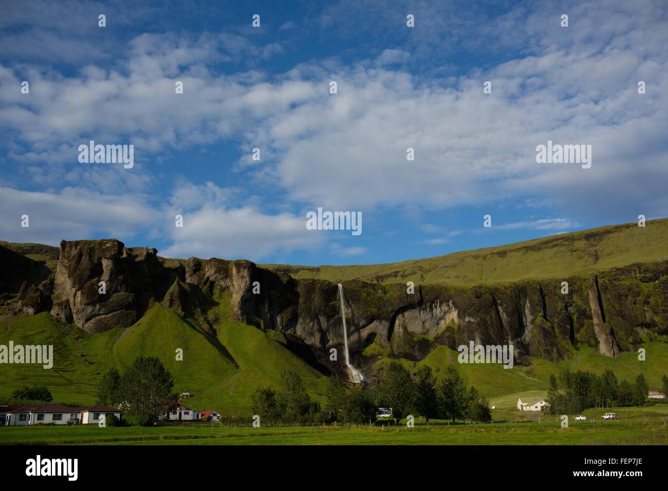 Remoto villaggio da una cascata che scorre oltre il verde lussureggiante mountain range, Islanda Foto Stock