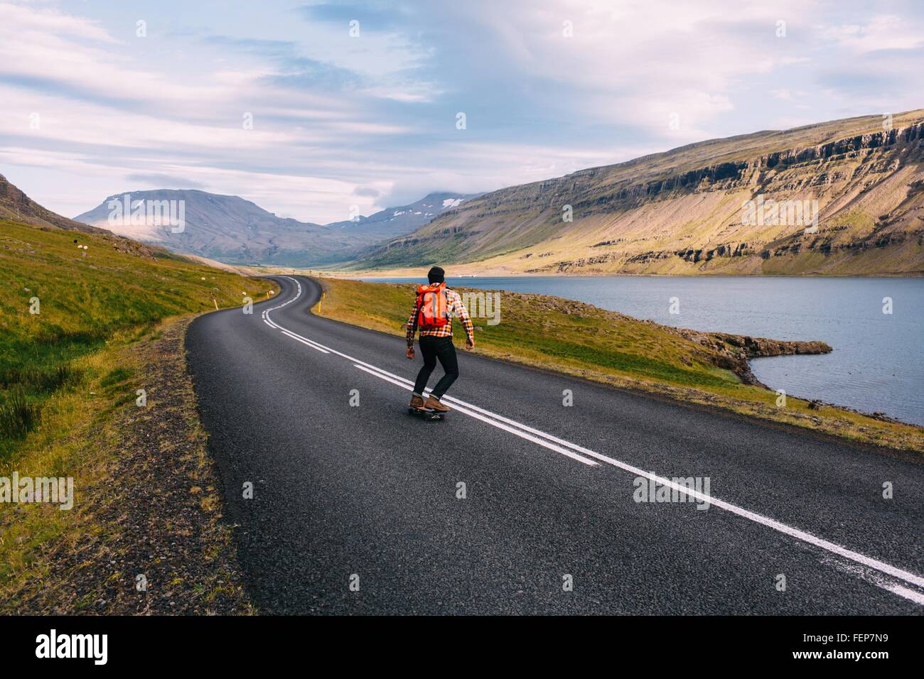 Vista posteriore della metà uomo adulto skateboarding curvando sulla strada aperta dal lago, Islanda Foto Stock