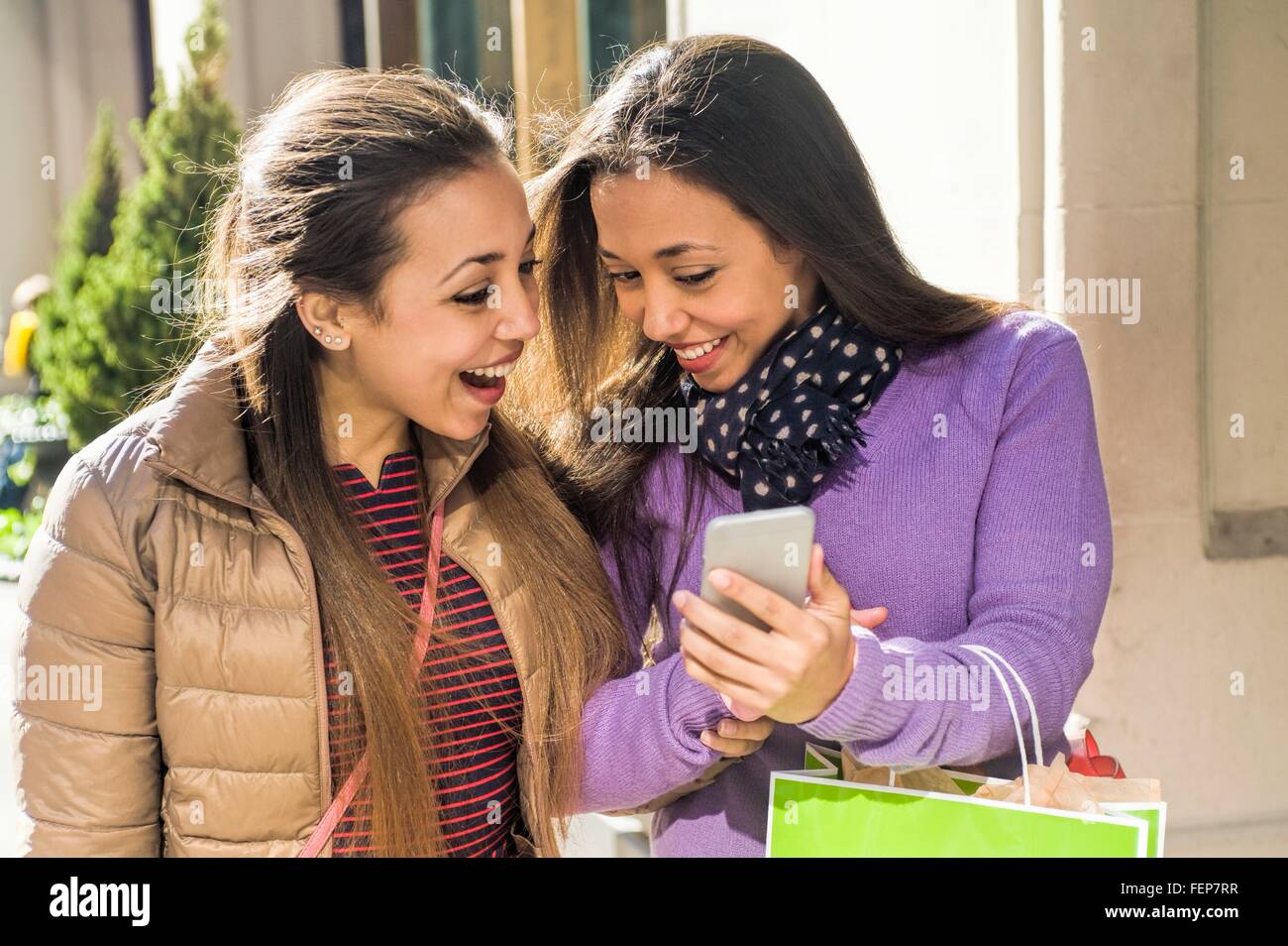 Giovani adulti femmine gemelli in città con le borse della spesa ridere del testo dello smartphone Foto Stock
