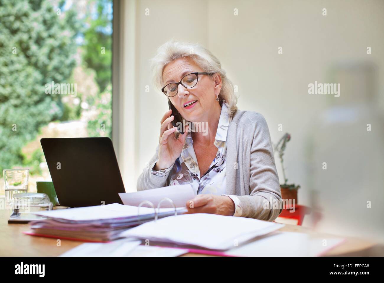 Senior donna seduta al tavolo, guardando attraverso documenti, utilizza lo smartphone Foto Stock