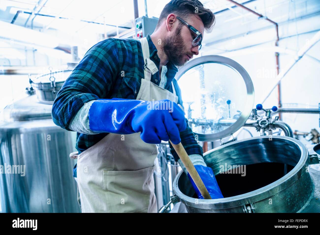 Vista laterale del giovane uomo in birreria indossando grembiuli e guanti protettivi di birra di test nel serbatoio Foto Stock