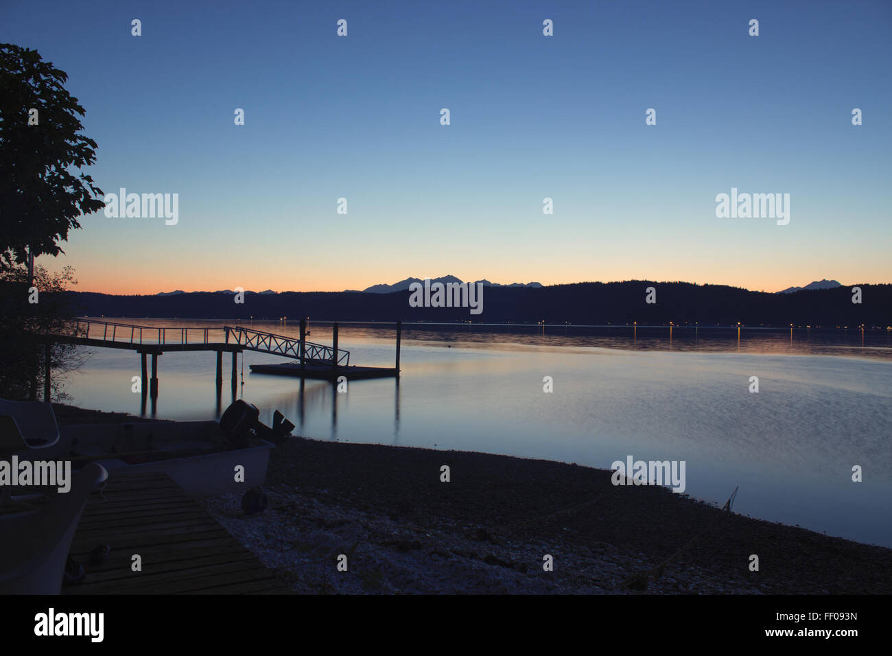Un molo si estende in un lago calmo, con riflessi della struttura e dell'ambiente circostante riflessi nell'acqua. La scena serena è impreziosita dal tramonto e dal cielo blu, creando un'atmosfera tranquilla all'aperto. Foto Stock
