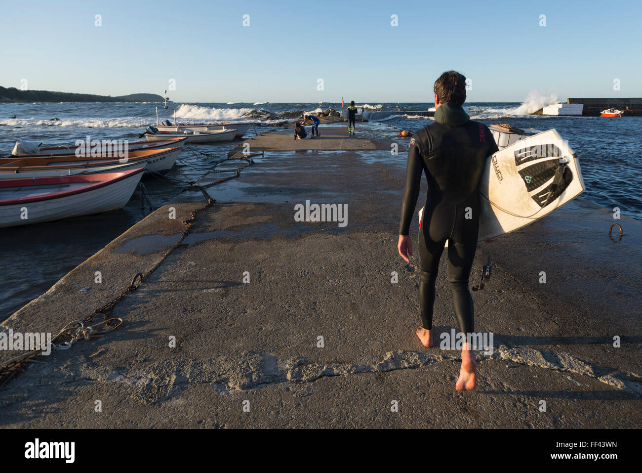 Un surfista maschio in wetsuit cammina verso il mare sulla mole del piccolo porto di pesca Vik su un giorno d'estate,costa orientale,Mar Baltico,Svezia Foto Stock