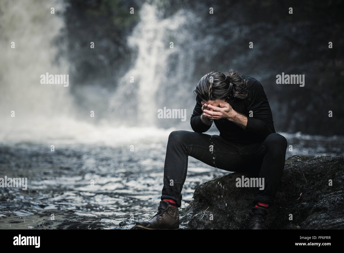 Un uomo seduto lavando il suo volto in un torrente di montagna da una cascata. Foto Stock