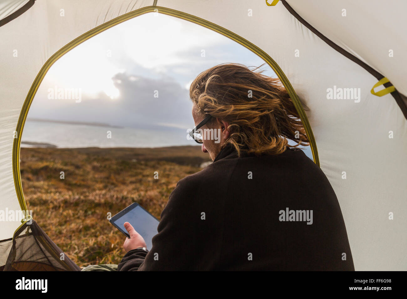 Un uomo seduto al riparo di una tenda guardando fuori, tenendo una tavoletta digitale Foto Stock