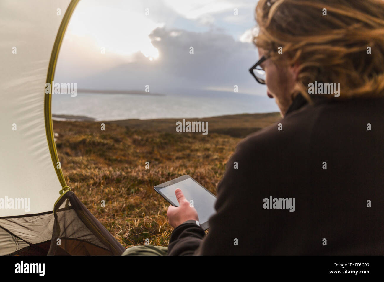 Un uomo seduto al riparo di una tenda guardando fuori, tenendo una tavoletta digitale Foto Stock
