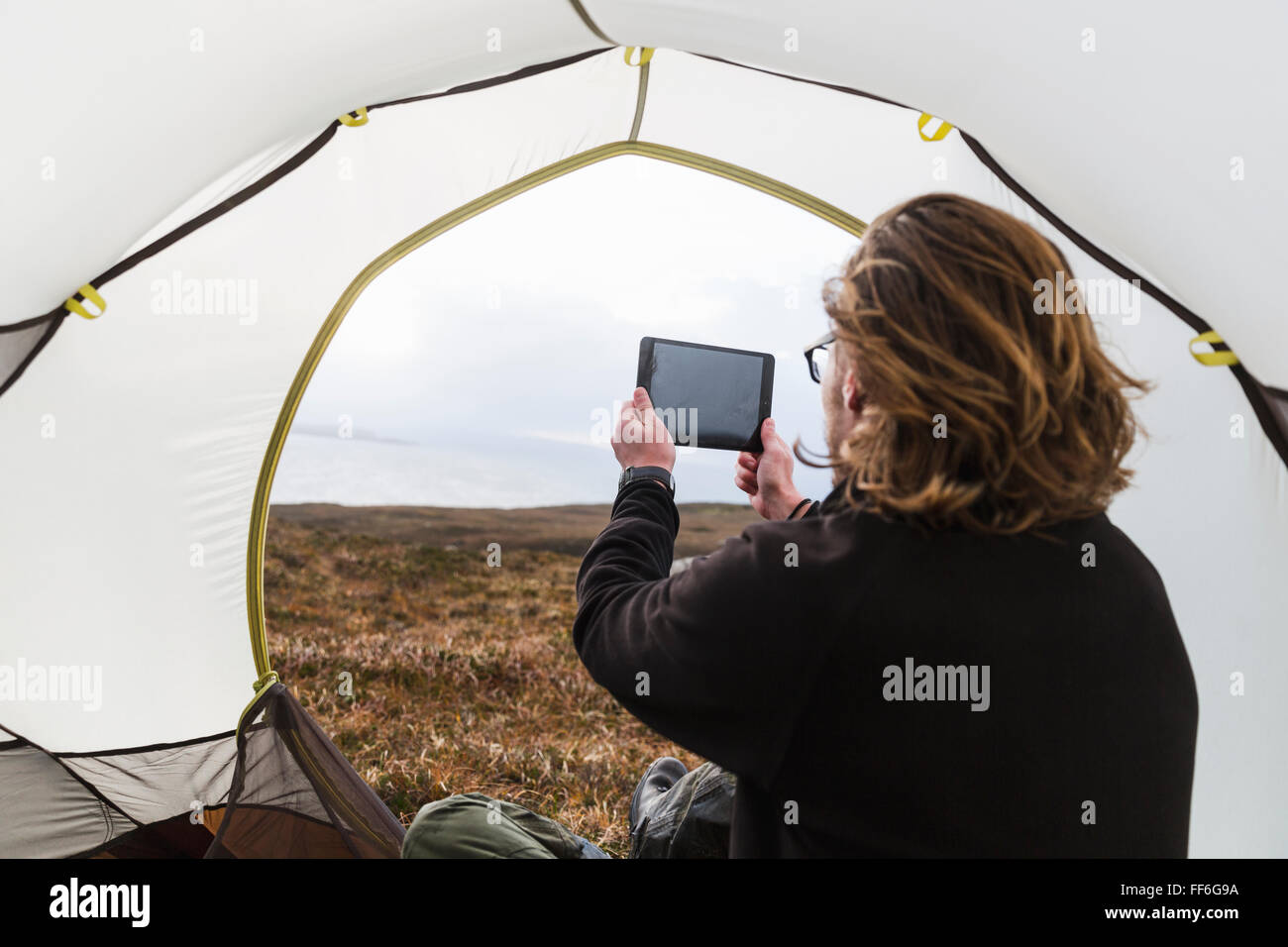 Un uomo seduto al riparo di una tenda guardando fuori, scattare una fotografia con una tavoletta digitale Foto Stock