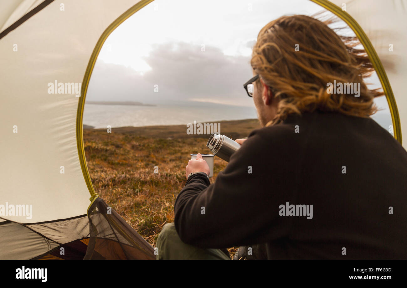 Un uomo seduto al riparo di una tenda guardando fuori il versamento di una bevanda a partire da una memoria flash. Campeggio selvaggio. Foto Stock