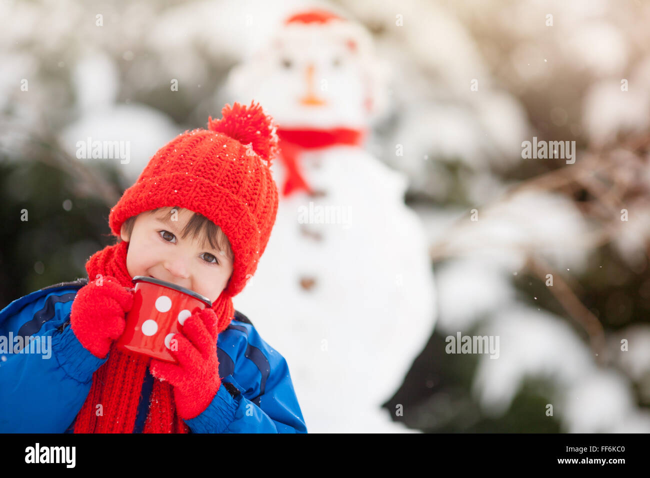 Felice bellissimo edificio bambino pupazzo di neve nel giardino invernale, tenendo tazza di tè caldo, sorridente in telecamera Foto Stock