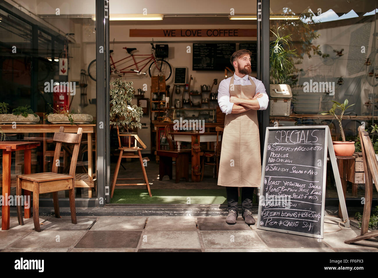 Ritratto di un giovane uomo in piedi nella parte anteriore della sua caffetteria. Giovane uomo con la barba di indossare un grembiule in piedi con le braccia cros Foto Stock