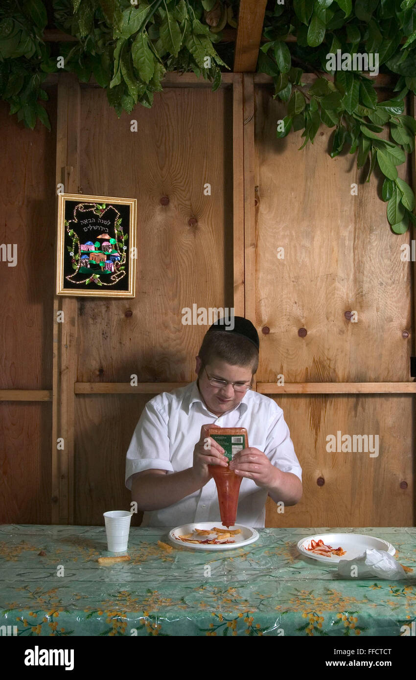 Boruch Shloima spremitura di salsa di pomodoro sulla sua take away chips mentre seduto nella famiglia Sukkah durante il festival di Sukkot, la festa dei tabernacoli. La vacanza commemora i quaranta anni di periodo durante il quale i figli d'Israele erano nel deserto. In onore dei figli di Israele nel deserto, gli uomini abitano in rifugi temporanei. Questo rifugio è chiamato un Sukkah ha almeno tre lati e un aprire parzialmente il tetto coperto di verde. Foto Stock