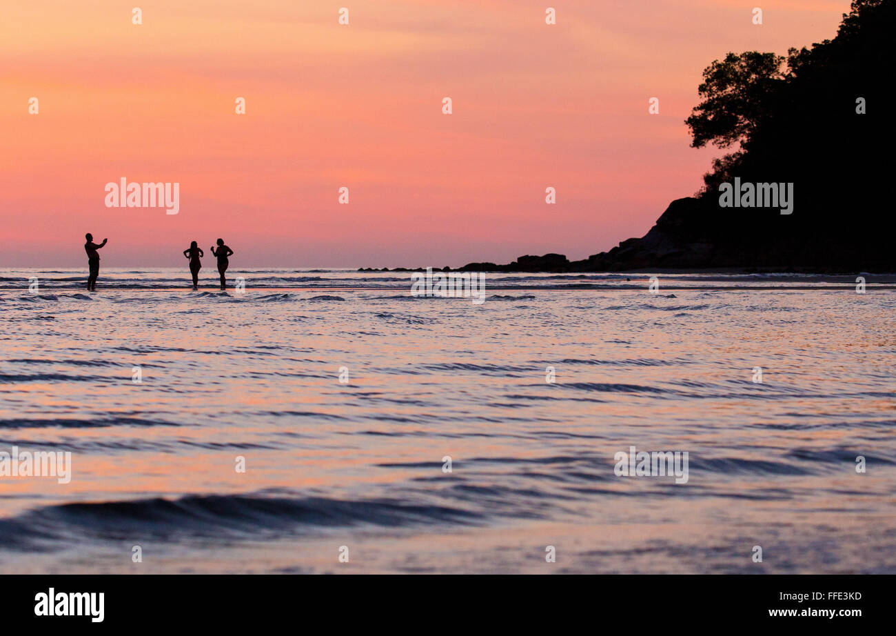 Tre persone in piedi in mare al tramonto off una spiaggia nella parte occidentale di Sabah, Malesia. Una persona che sta prendendo una foto del tramonto. Foto Stock
