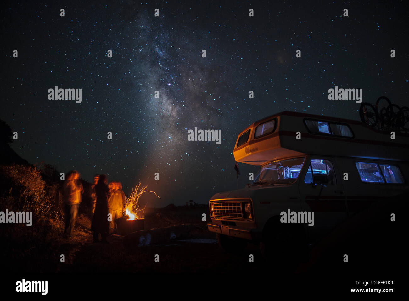 Un gruppo di viaggiatori stanno intorno a un falò e le scintille volano in alto verso la Via Lattea sopra di loro. Il cielo stellato è luminoso a Na Foto Stock