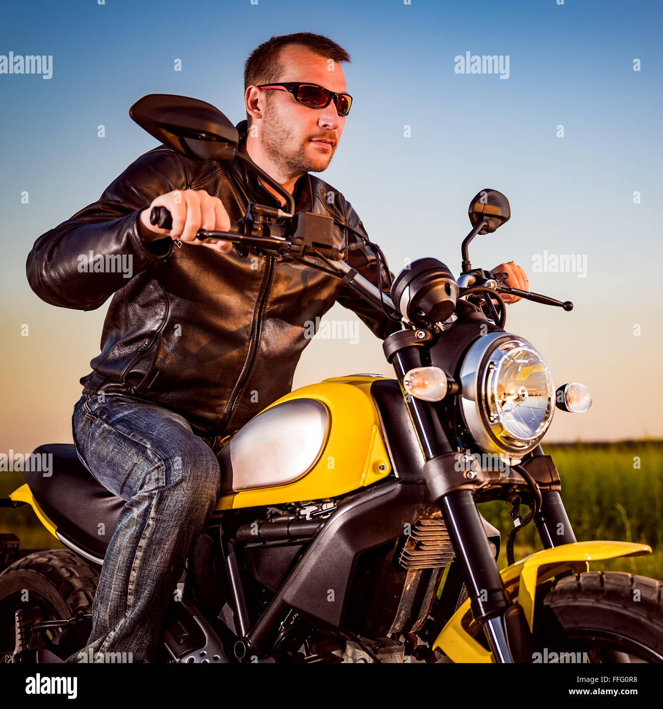 Biker uomo che indossa una giacca di pelle e occhiali da sole seduto sulla sua moto guardando il tramonto. Foto Stock Biker uomo che indossa una giacca di pelle e occhiali da sole seduto sulla sua moto guardando il tramonto. Foto Stock