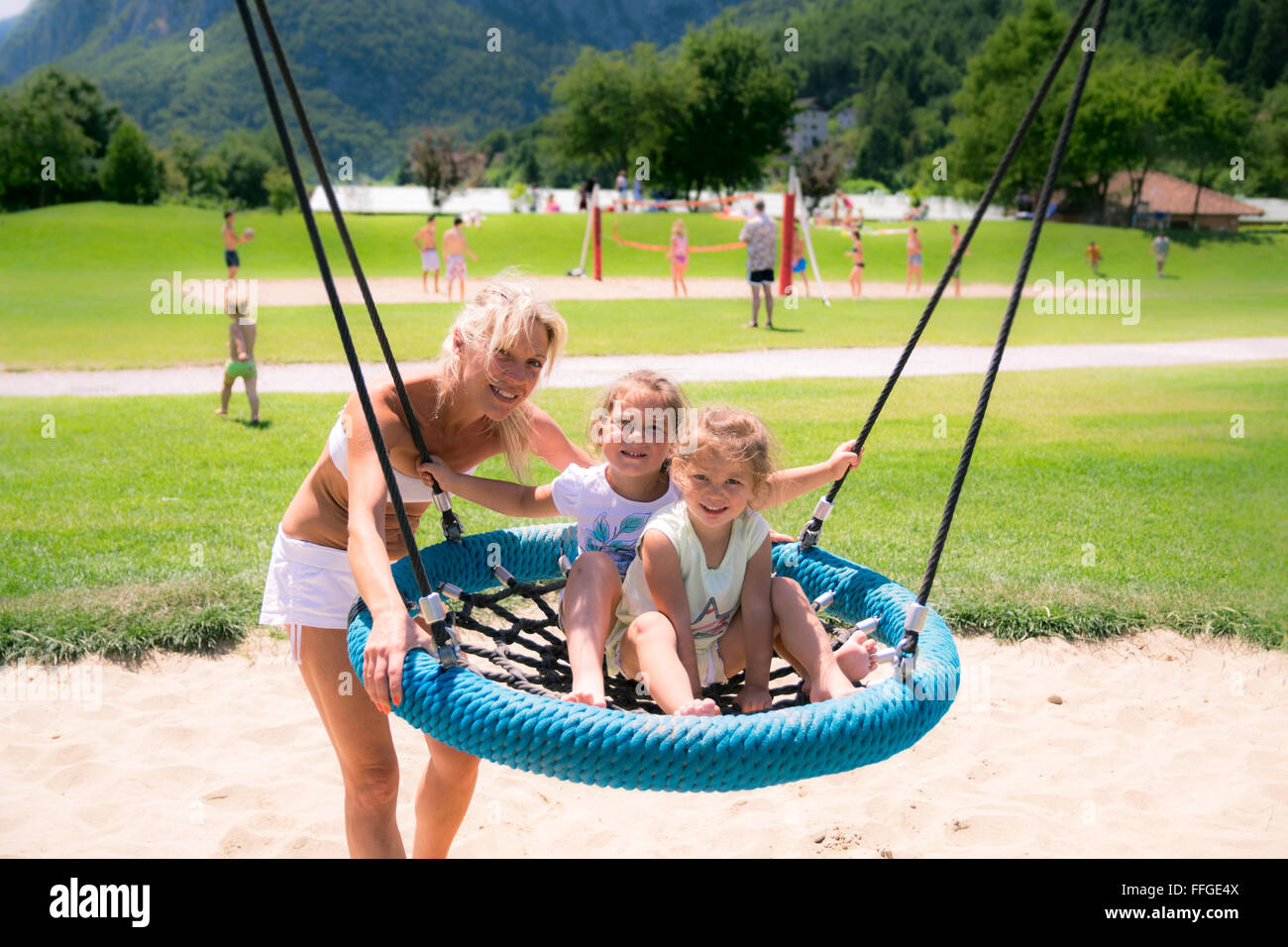 Blonde mom spinge le ragazze sul round swing corda in un parco giochi. Foto Stock