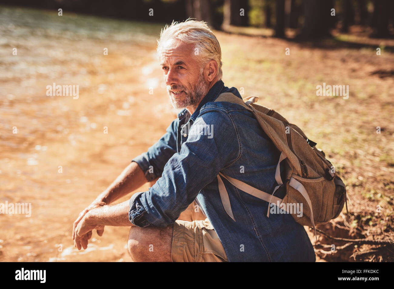 Ritratto di senior escursionista maschile seduto accanto a un lago e guardando lontano. Uomo maturo prendendo una pausa dalla sua escursione e guardando th Foto Stock