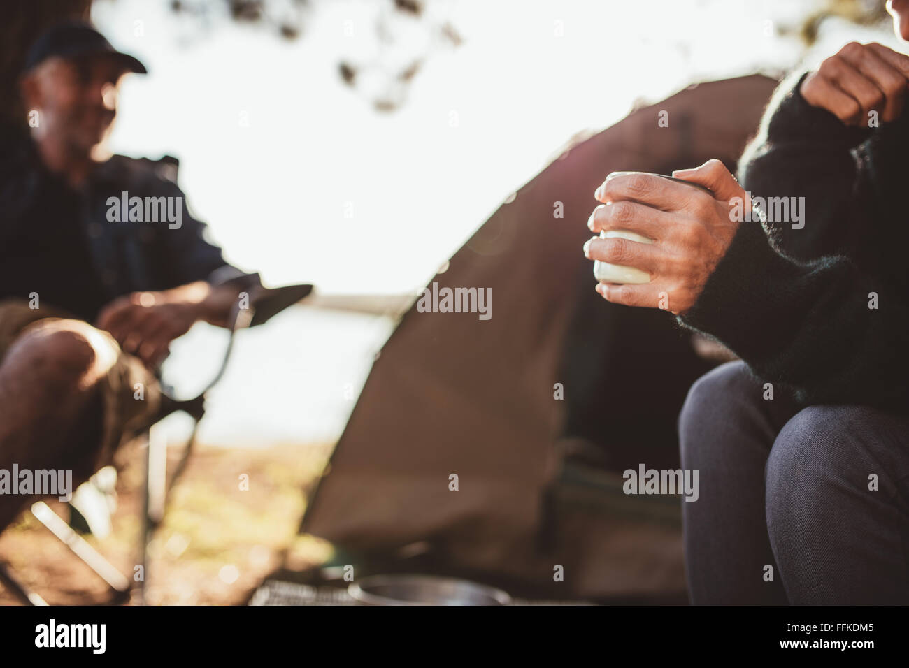 Close up ritratto di una mano di una donna azienda caffè al campeggio con un uomo in background, sia la seduta al di fuori della tenda. Foto Stock