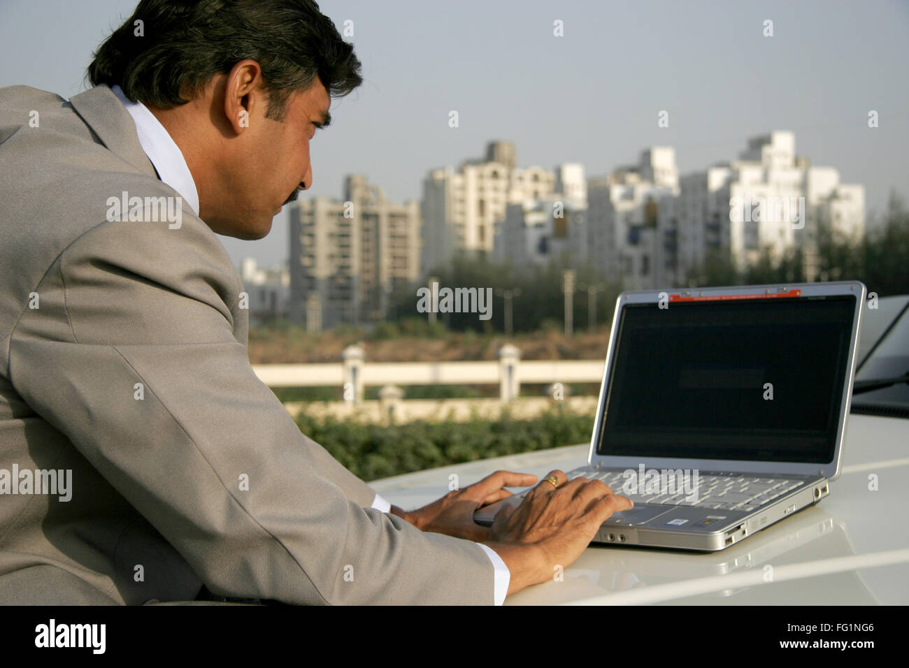 Commerciante indiano in cappotto grigio e nero pant utilizzando laptop cofano auto contro alcuni edifici aziendali Pune City Maharashtra Foto Stock