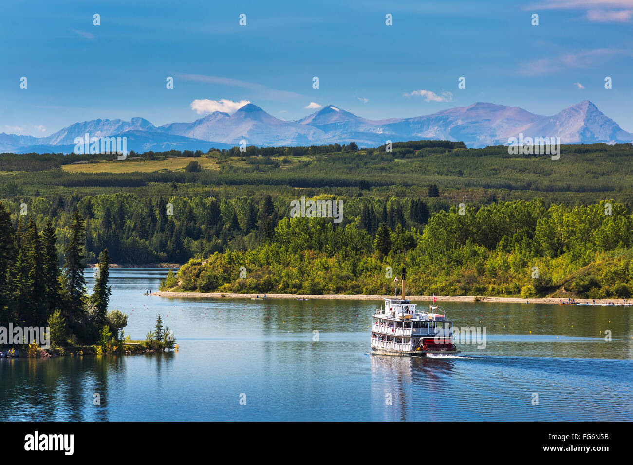 Pedalo' barca sul lago con viale alberato costa, colline e montagne sullo sfondo e il cielo blu e nuvole; Calgary, Alberta, Canada Foto Stock