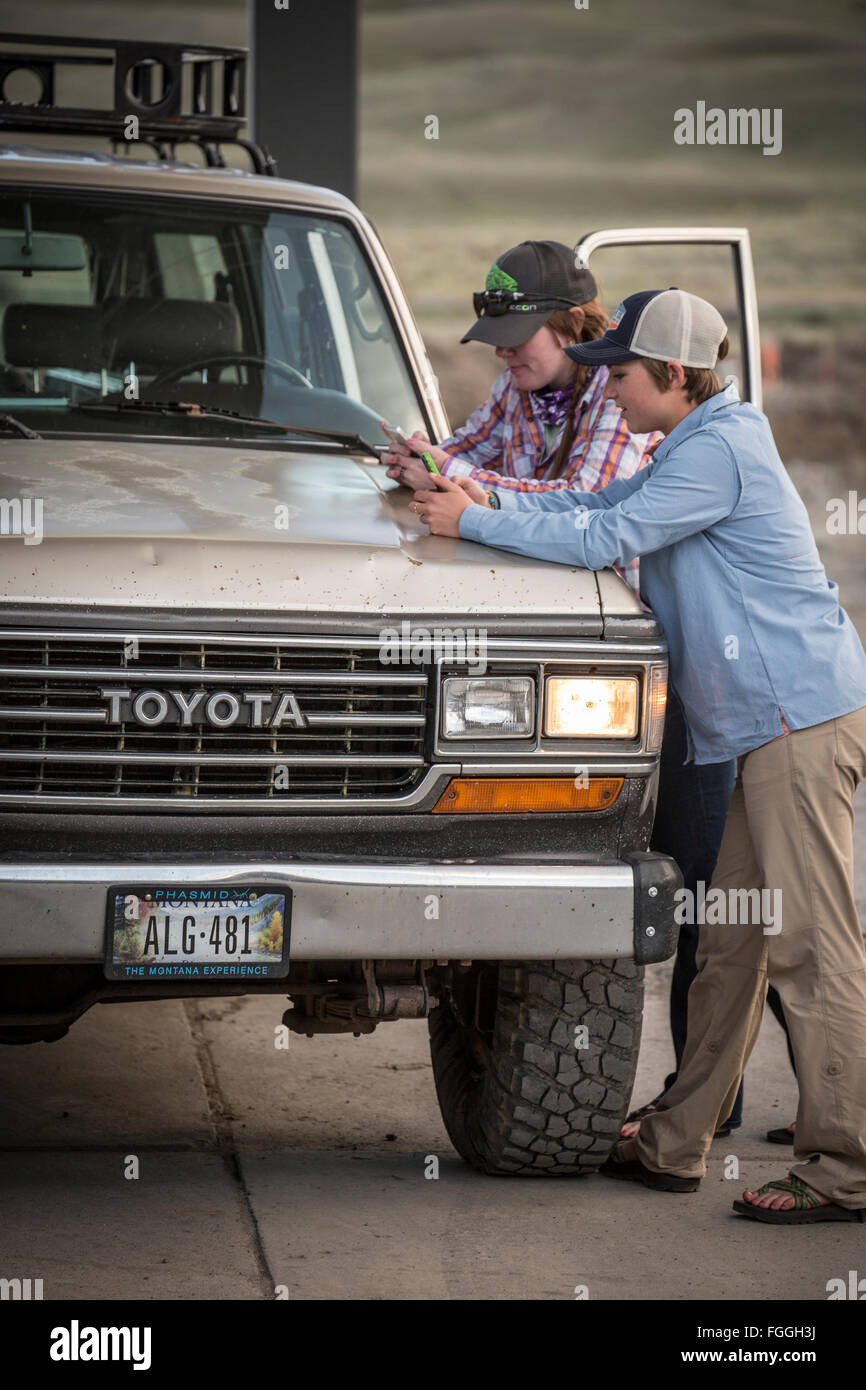 Due ragazze testo sui loro telefoni mentre in corrispondenza di una zona rurale montana stazione di gas con un 1990 Toyota Land Cruiser. Foto Stock