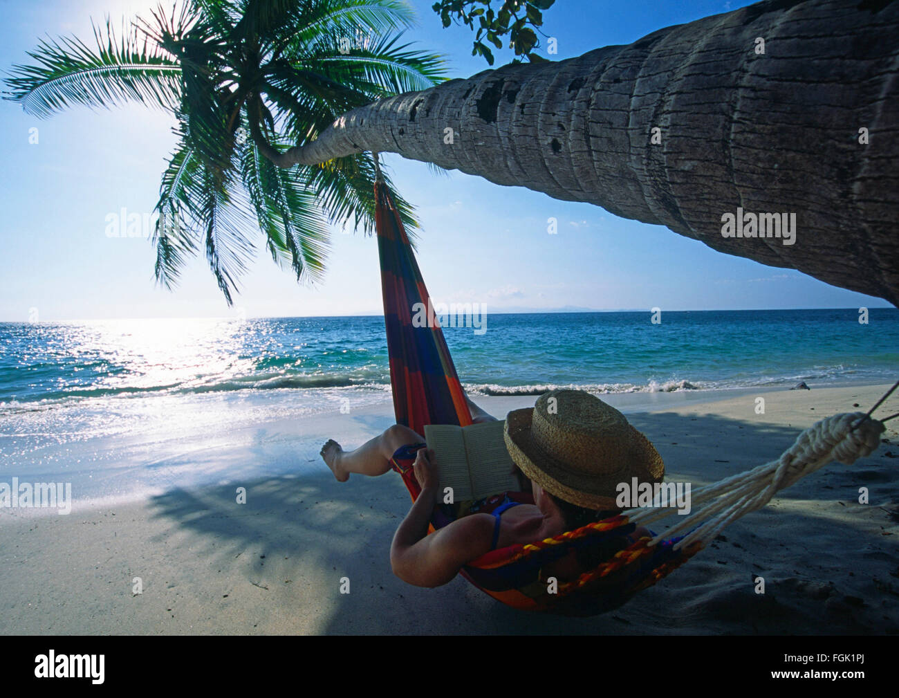 Una donna in un amaca a leggere un libro sotto una palma da cocco albero piegato sopra l'acqua su una spiaggia sulla costa del Pacifico di Costa Rica Foto Stock