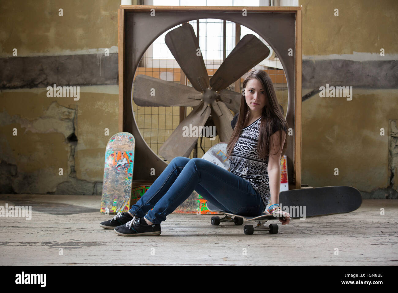 Bella bruna donna seduta su uno skateboard di fronte un grande ventilatore industriale Foto Stock
