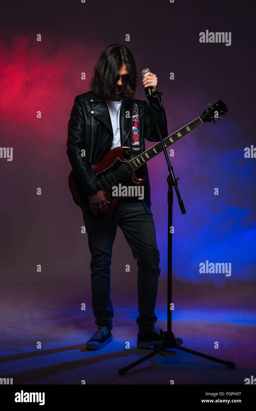 Bel giovane cantante maschio con i capelli lunghi a cantare nel microfono e suonare la chitarra su colorato sfondo affumicato Foto Stock