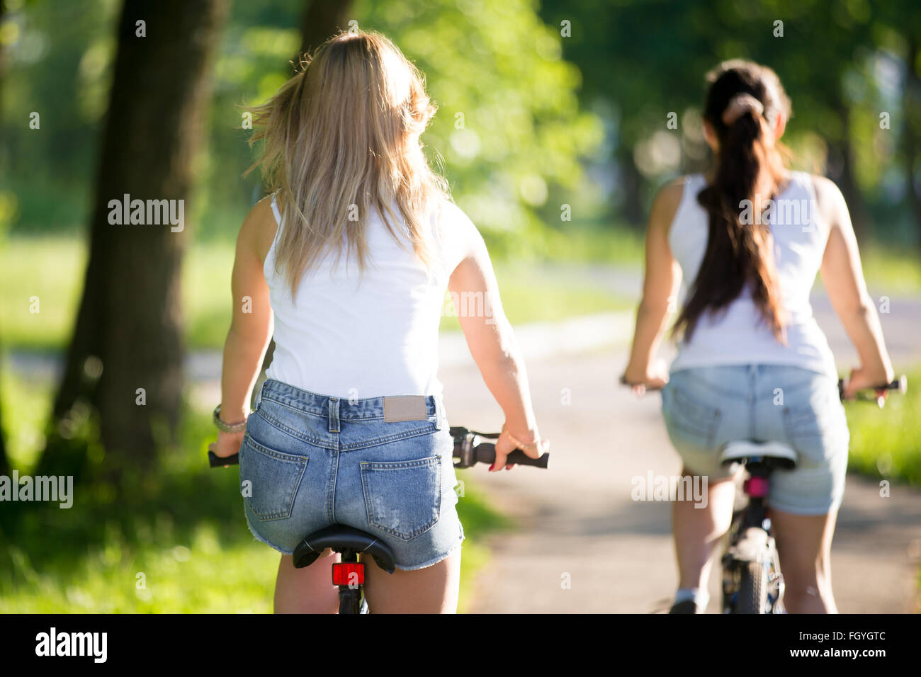 Due giovani donne belle ragazze indossano jeans corti mountain bike sul marciapiede sulla strada sulla soleggiata giornata estiva, vista posteriore Foto Stock