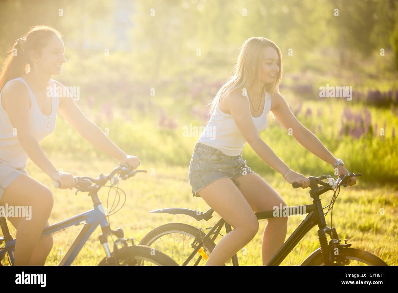 Due simpatici giovani sorridenti donne belle ragazze indossano jeans corti moto di equitazione nel parco in condizioni di luce solare intensa in estate Foto Stock