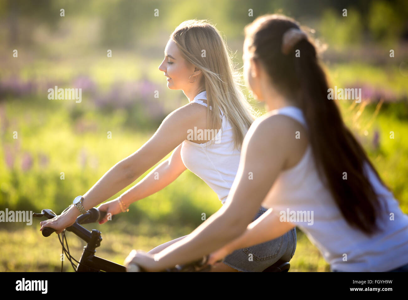 Due simpatici giovani sorridenti belle ragazze che indossano bianco casual canotte godere di escursioni in bicicletta nel parco in condizioni di luce solare intensa Foto Stock
