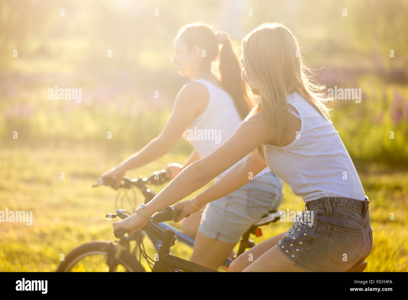 Due giovani graziosi felice belle ragazze che indossano bianco casual canotte e gli shorts in denim biciclette equitazione nel parco in bright Foto Stock