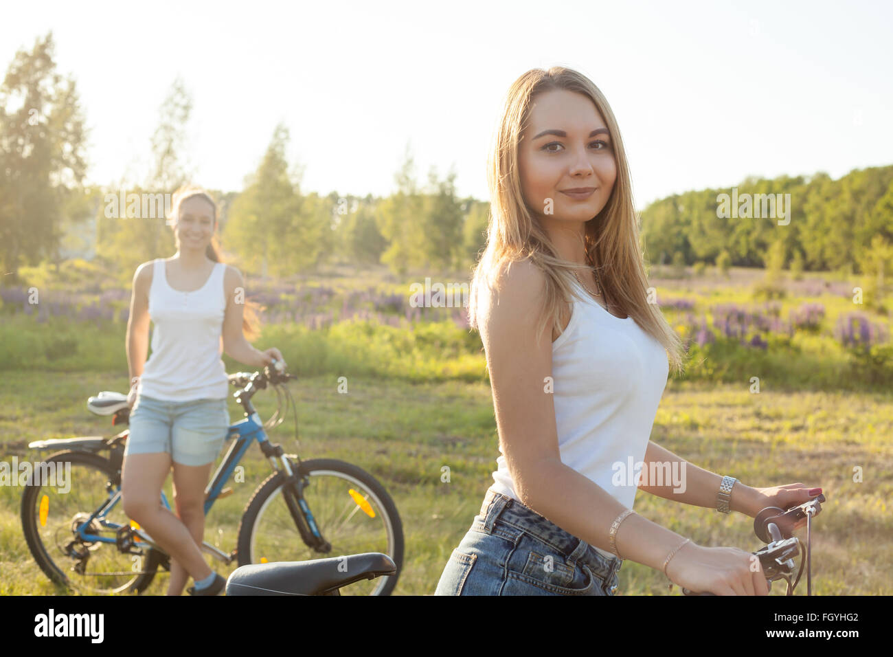 Due belle ciclista fidanzate indossando bianco casual canotte e jeans corti in piedi con la moto in campagna parco Foto Stock