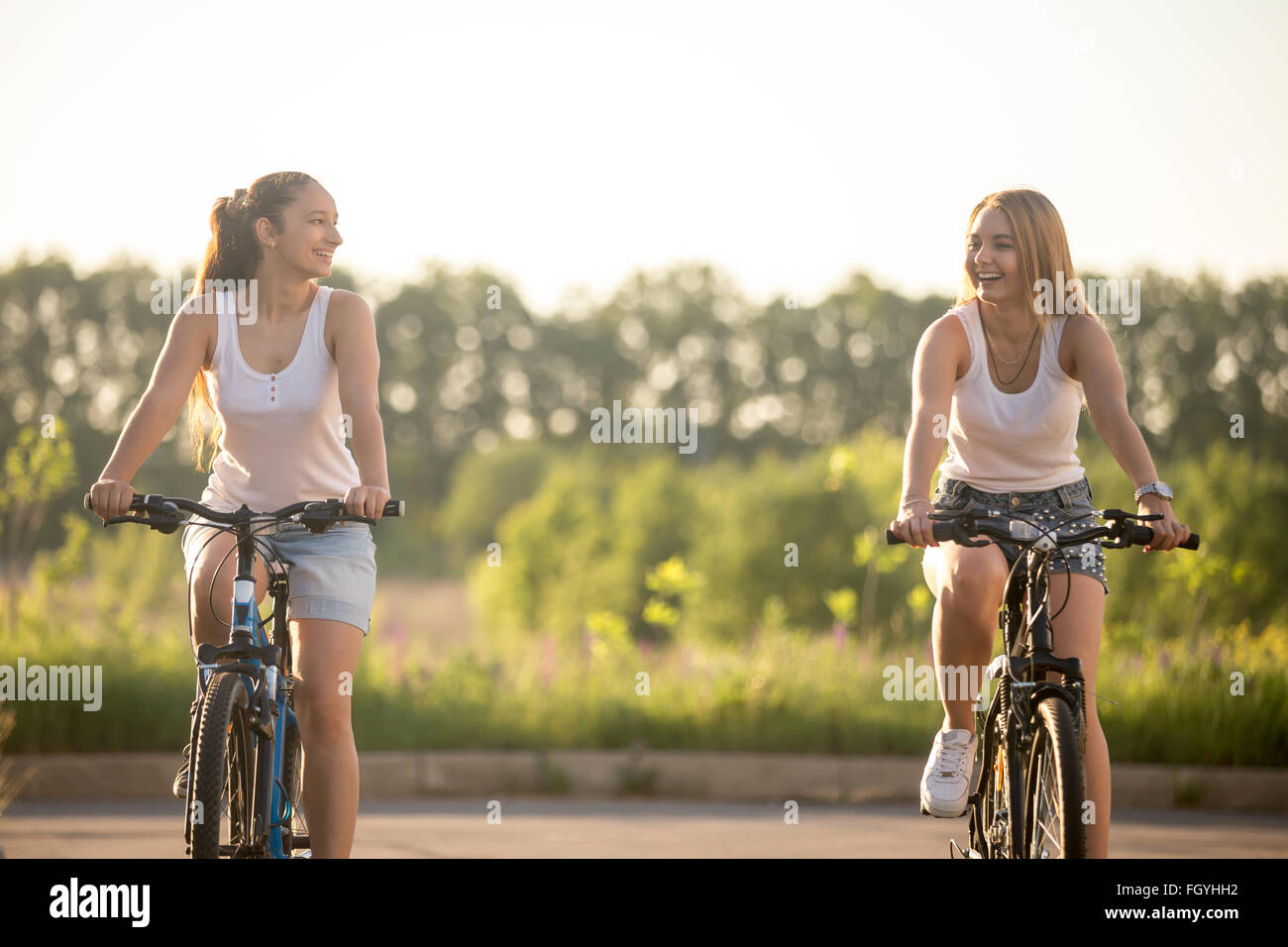 Due belle giovani donne equitazione biciclette indossando bianco casual canotte e jeans corti in parco sulle soleggiate giornate estive Foto Stock