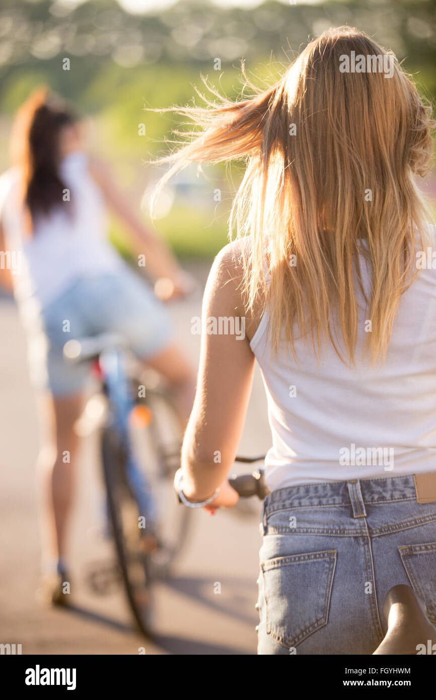 Vista posteriore di due belle ragazze adolescenti a partire bike ride indossando bianco casual canotte e jeans corti sul park road Foto Stock